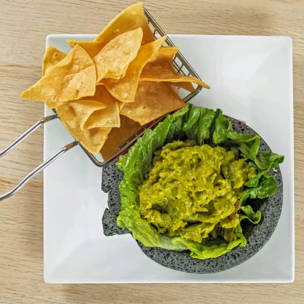 Guacamole in a dark bowl with lettuce, served with a basket of tortilla chips on a white plate.