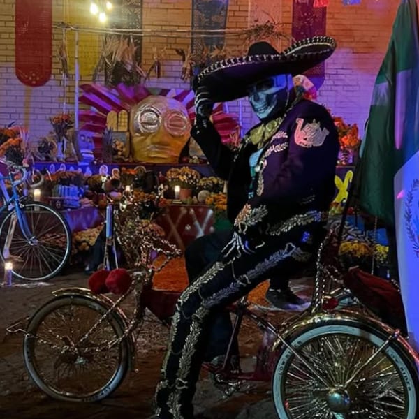 Day of the Dead skeleton figure in mariachi attire on a gold bicycle, surrounded by offerings.