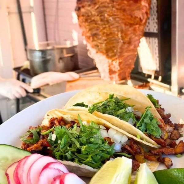 Al pastor tacos with cilantro, onions, limes, and radishes, with a vertical rotisserie in the background.