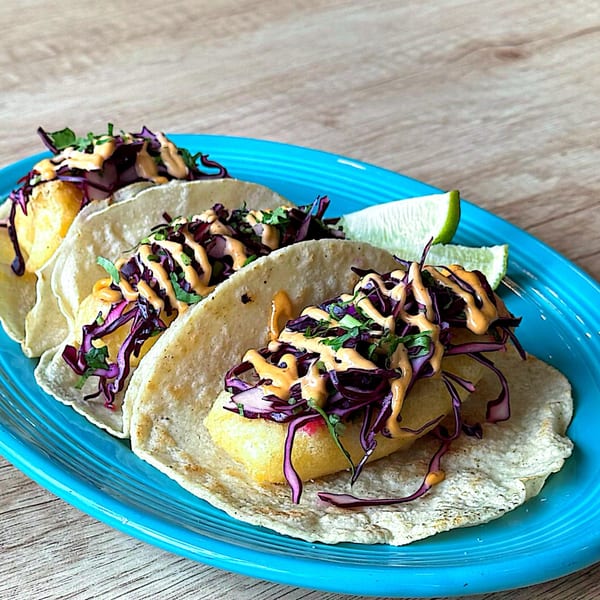 Three La Ingrata Camden fish tacos with purple cabbage, sauce, and lime on a blue plate.